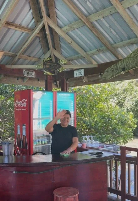 Person standing behind a wooden bar under a pavilion, smiling and touching their head, with Coca-Cola fridge and green trees in the background.