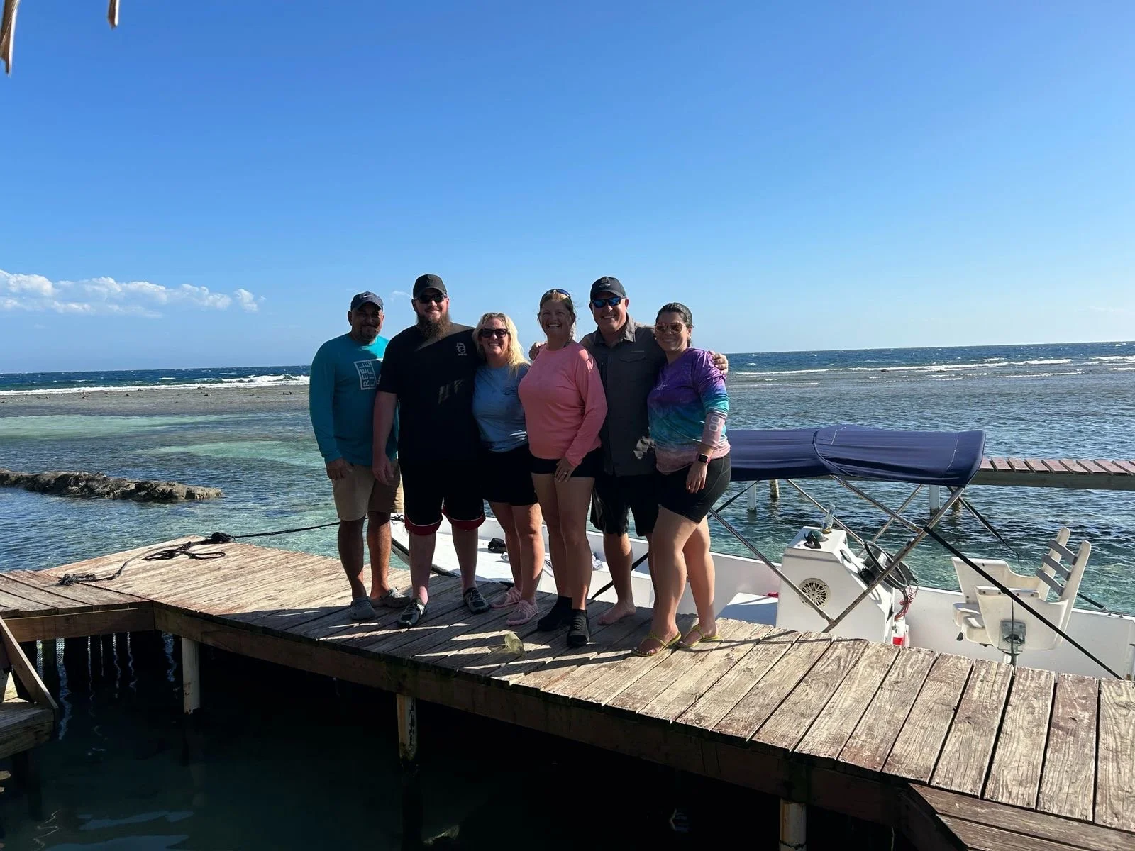 Group of six people standing on a wooden dock by the water, with a boat in the background, on a sunny day with blue sky and ocean waves.