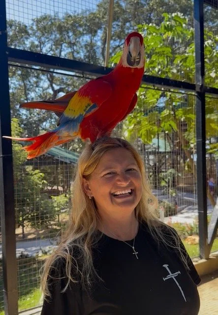 Woman smiling at a colorful parrot perched above her head inside a bird enclosure.