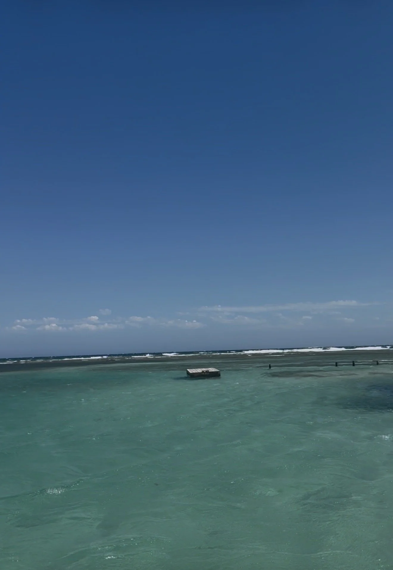 A calm ocean with turquoise water in the foreground, a broader ocean with small waves in the background, and a clear blue sky with a few clouds.