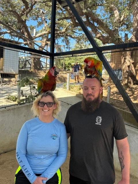 A woman and a man standing inside an outdoor aviary with two parrots perched on their heads. The woman is smiling and wearing glasses, while the man has a beard and a neutral expression.