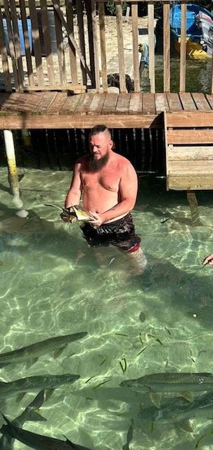 A man is standing in clear water by a wooden dock, holding a fish he caught. There are multiple fish swimming nearby in the water.