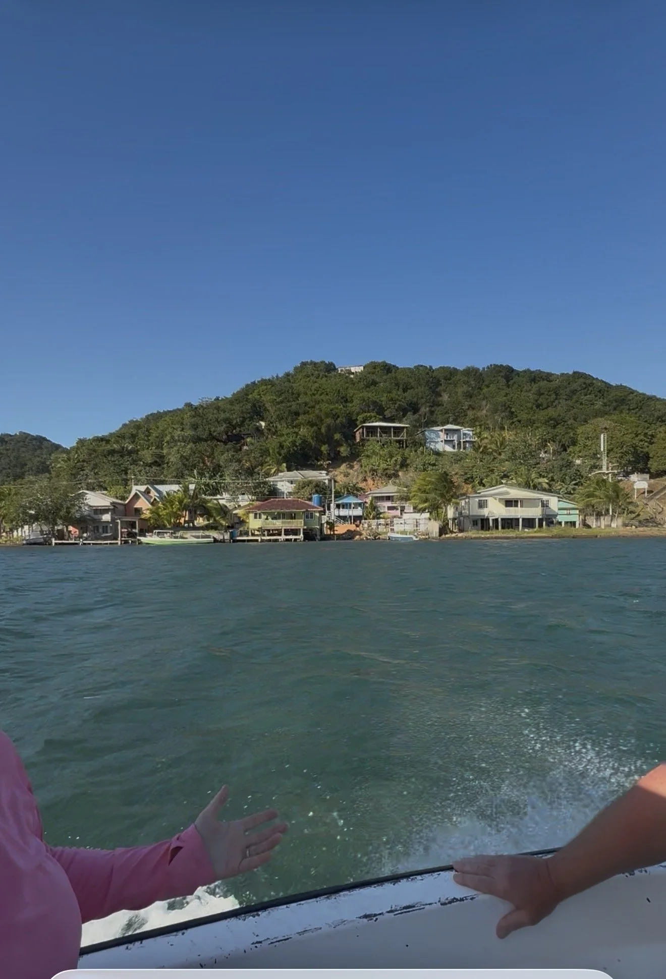 View of waterfront houses along the shore with hills in the background, as seen from a boat on the water.