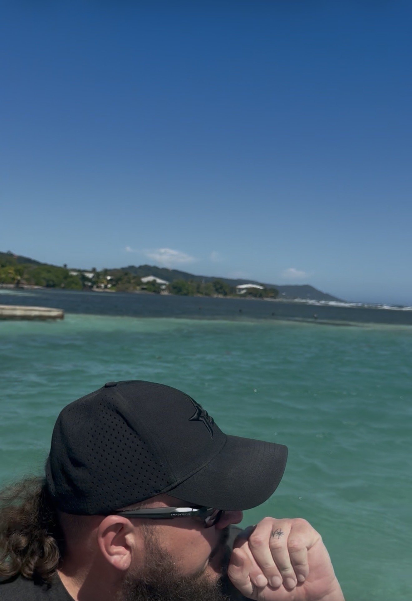 A man with a beard wearing a black cap and glasses, sitting by a turquoise body of water on a sunny day, with hills and houses in the background.