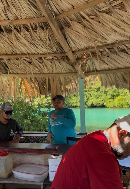 Group of people under a thatched roof, with a green and water background, possibly at a tropical outdoor setting