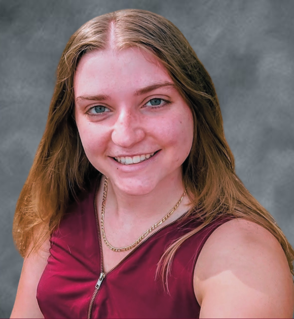 A young woman with long, light brown hair and blue eyes, smiling, wearing a burgundy sleeveless top and a gold chain necklace, against a gray background.