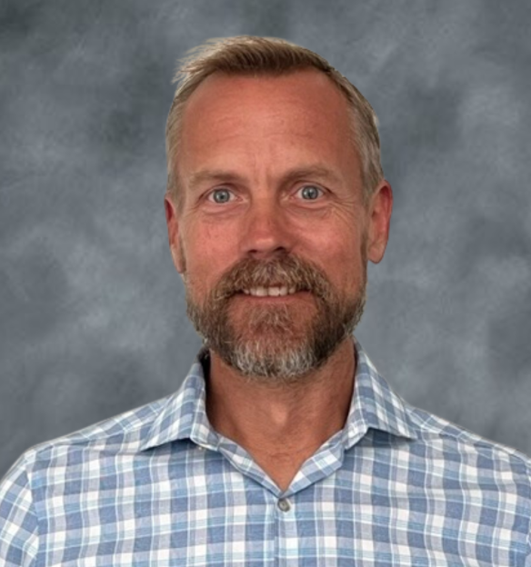 Portrait of a middle-aged man with light skin, short blonde hair, a beard, and blue eyes, wearing a blue and white checkered shirt, standing against a gray background.