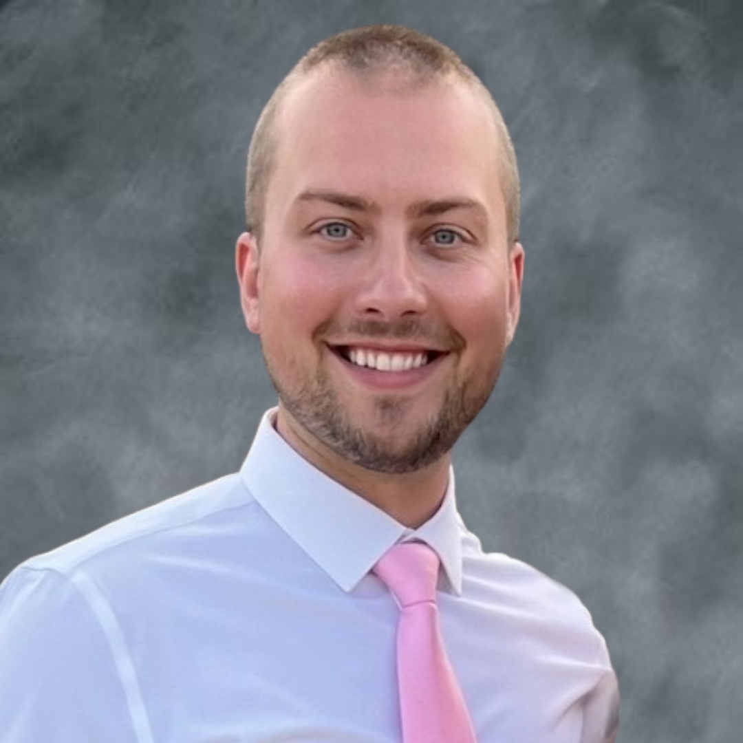 A smiling man with short hair, wearing a white shirt and pink tie, against a neutral gray background.