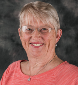 Portrait of an elderly woman with short gray hair, wearing glasses and a coral-colored top, smiling at the camera.