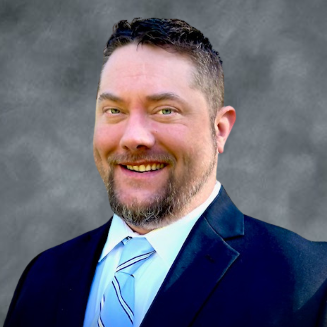 A man with light skin, short dark hair, and a beard, wearing a black suit, white shirt, and striped tie, smiling in front of a gray background.