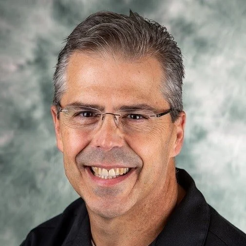 A smiling middle-aged man with glasses, short gray hair, wearing a black shirt, in front of a green and gray mottled background.