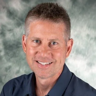 A smiling man with short gray hair, wearing a dark blue shirt, against a green and gray mottled background.