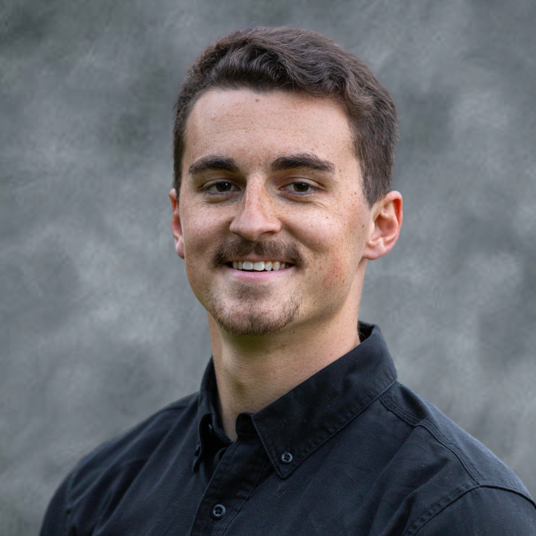 A young man with short dark hair, light skin, and a mustache, wearing a black button-up shirt, smiling outdoors with a blurred natural background.
