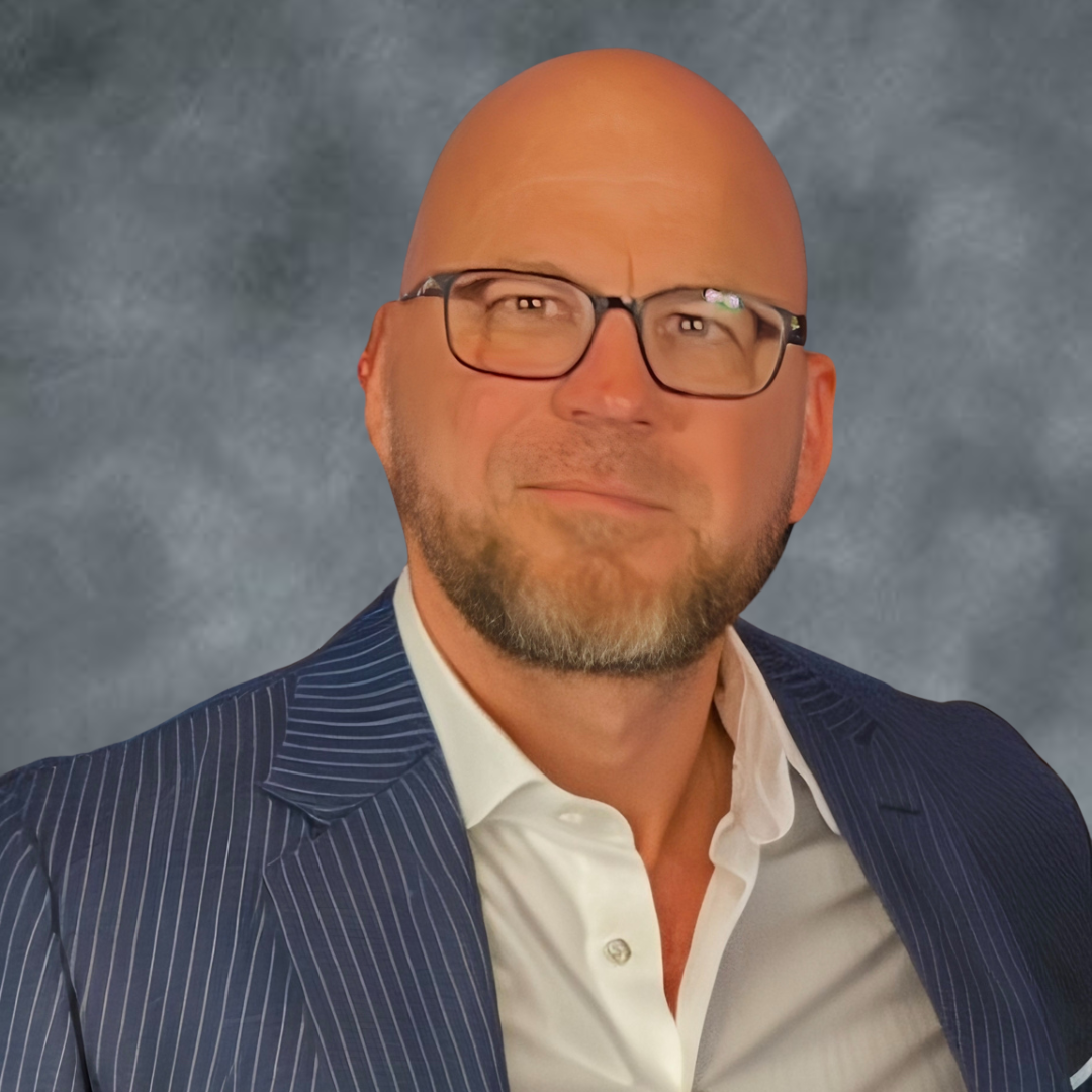 Portrait of a bald man with glasses, beard, wearing a dark striped suit and white shirt, against a gray textured background.
