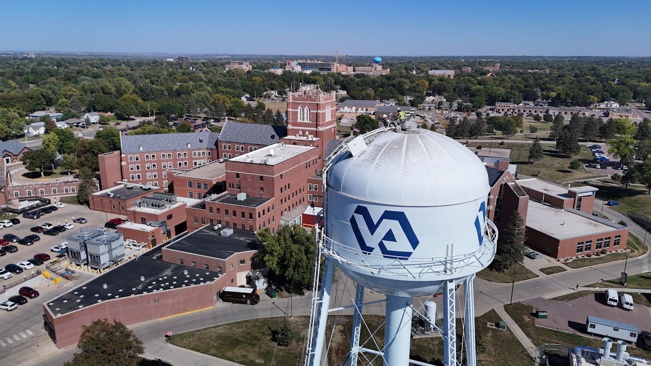Aerial view of a cityscape with a large water tower featuring the VA logo, surrounded by brick buildings, parking lots, and green trees.
