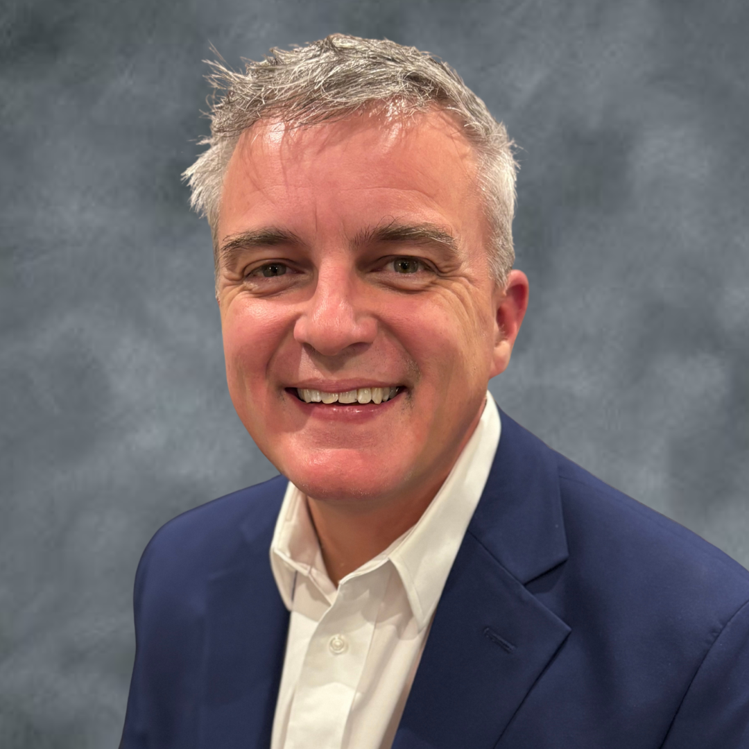 Headshot of a smiling middle-aged man with gray hair, wearing a white shirt and navy blazer, against a gray background.