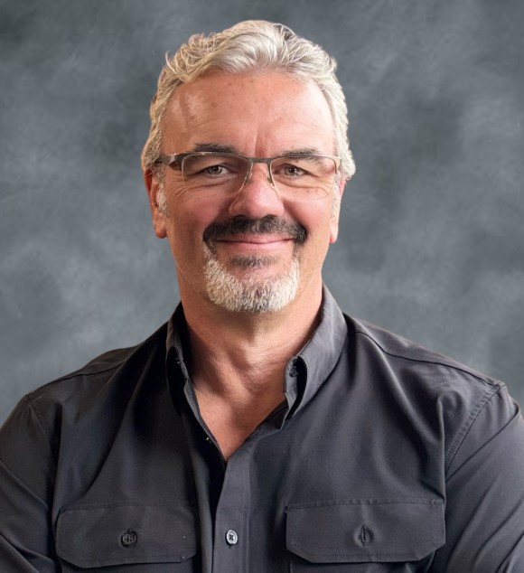 A middle-aged man with gray hair, glasses, and a beard smiling in front of a plain gray background.