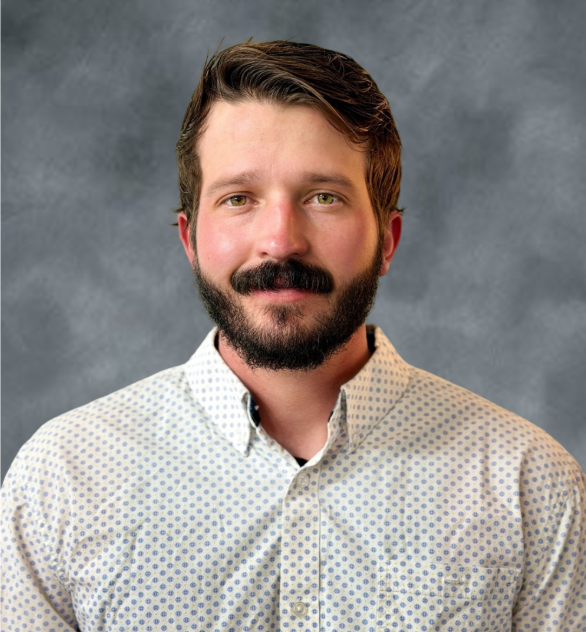 A man with short brown hair, a full beard and mustache, wearing a white dress shirt with small blue dots, standing against a gray background.