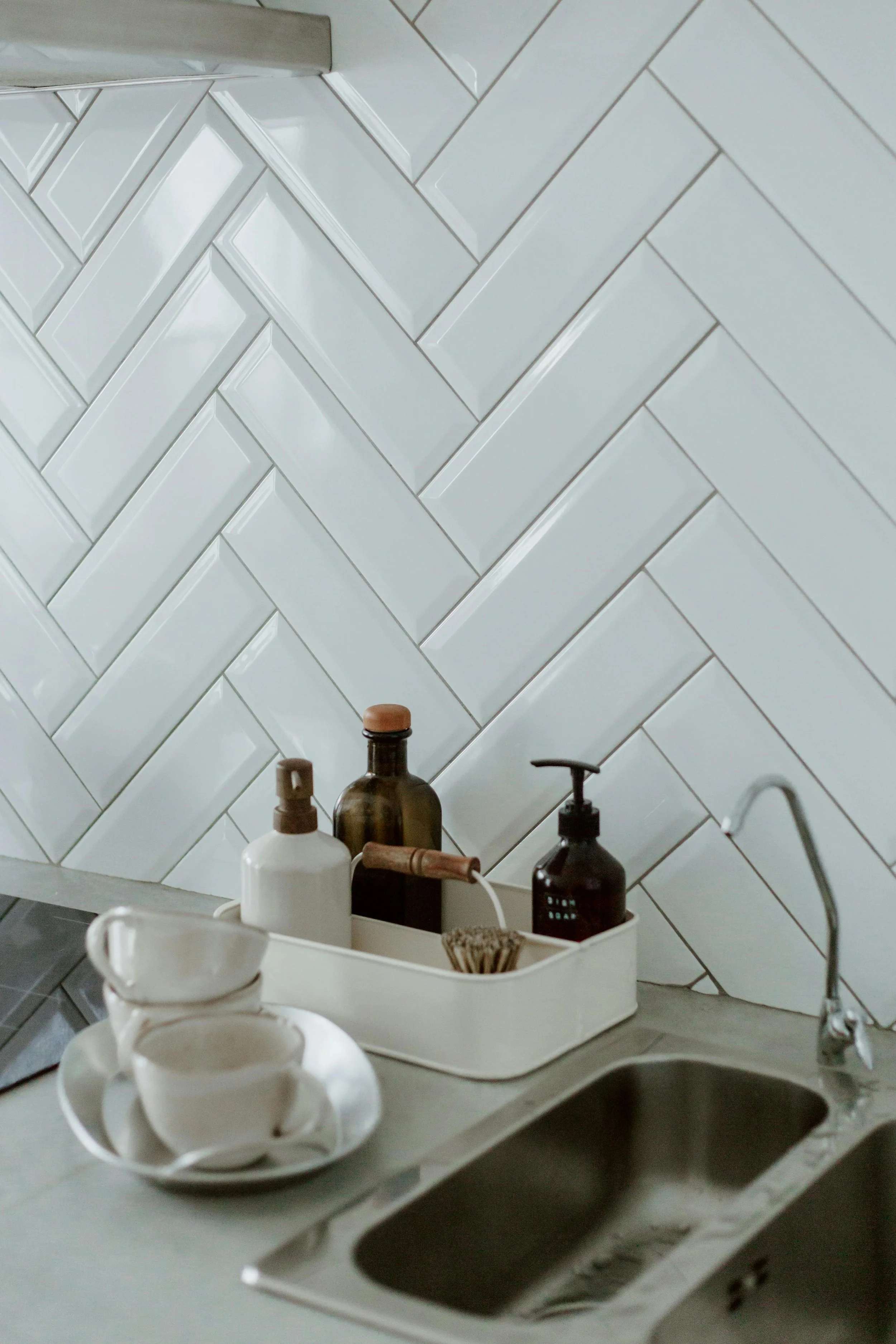 Kitchen counter with white dishes and bottles, white tile backsplash with a herringbone pattern, and a sink.