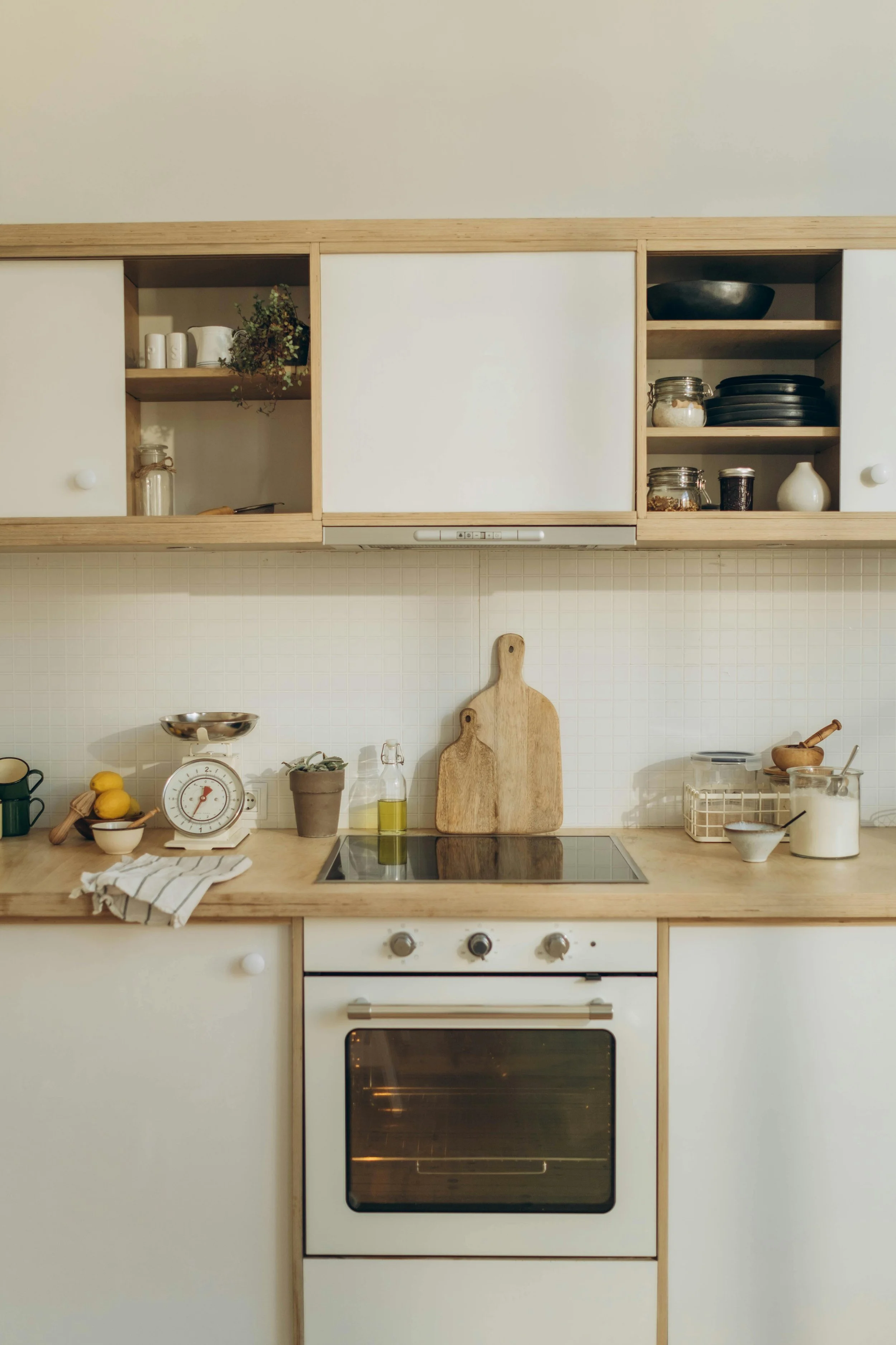 A minimalist kitchen with white cabinets, wooden countertops, and open shelving. The stove and oven are central, with kitchen utensils, jars, and a scale on the counter. Cutting boards lean against the backsplash.