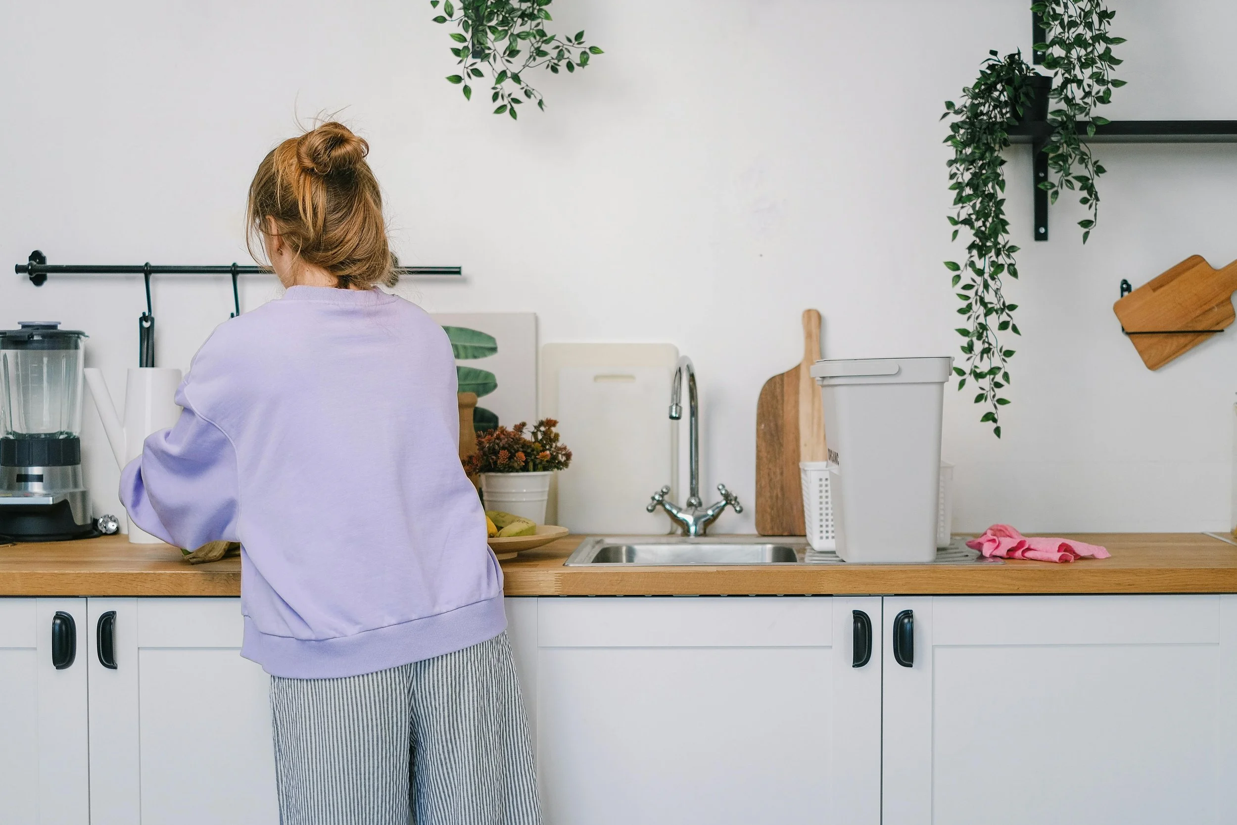 Person with red hair wearing a purple sweatshirt and striped pants standing at a kitchen counter, preparing food or cleaning, with green plants and kitchen tools in the background.