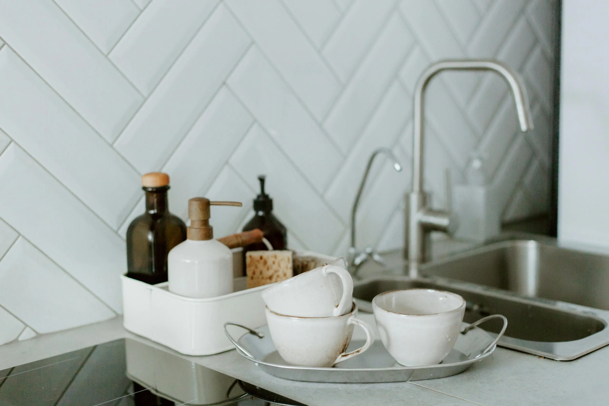 Kitchen sink area with three stacked white ceramic cups on a tray, soap dispensers, a sponge, and a modern stainless steel faucet against a herringbone tile backsplash.