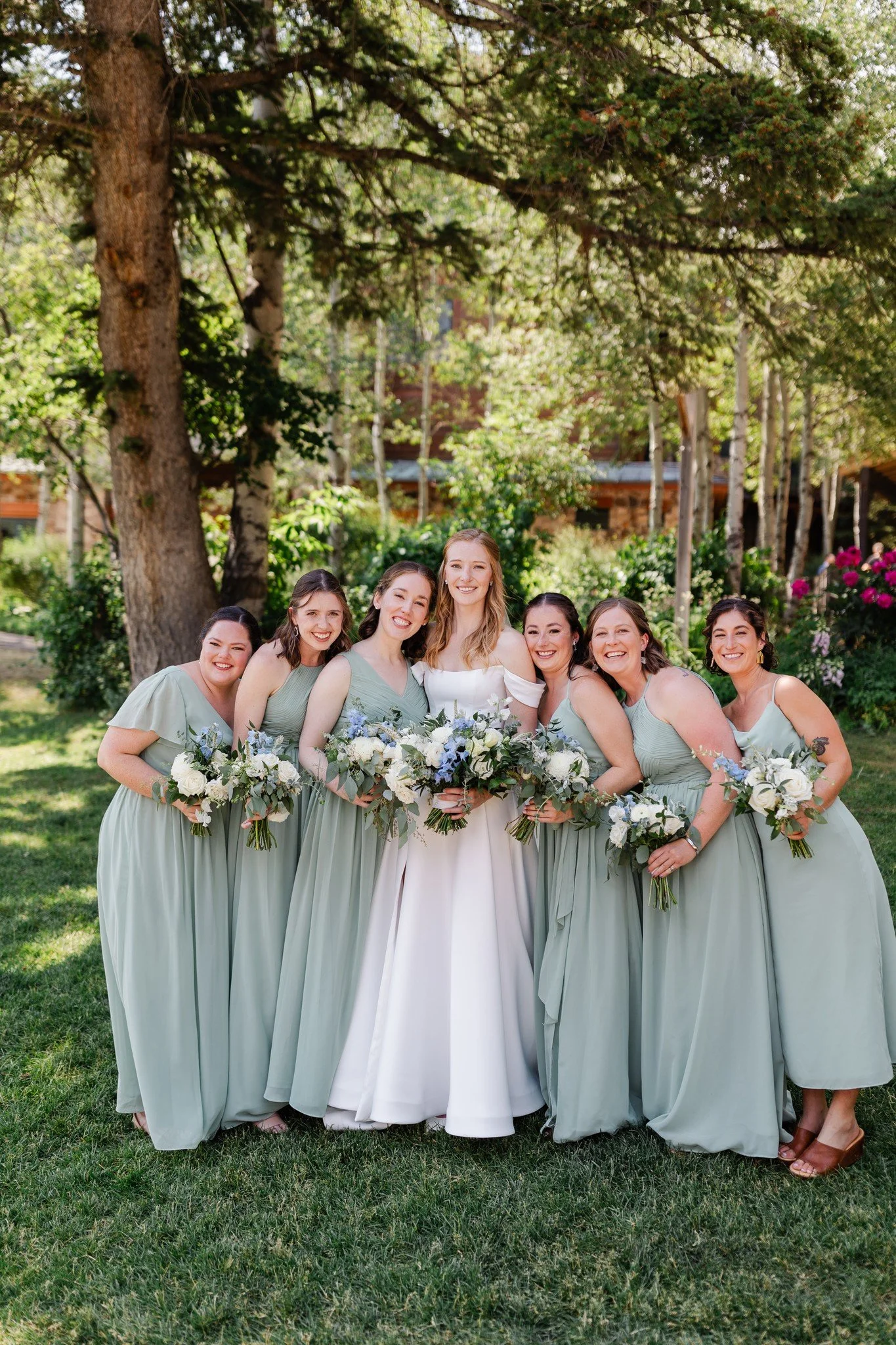 Group of seven women in pastel green bridesmaid dresses and one woman in a white wedding dress standing outdoors under large trees and smiling, holding bouquets of white and blue flowers during a wedding celebration.