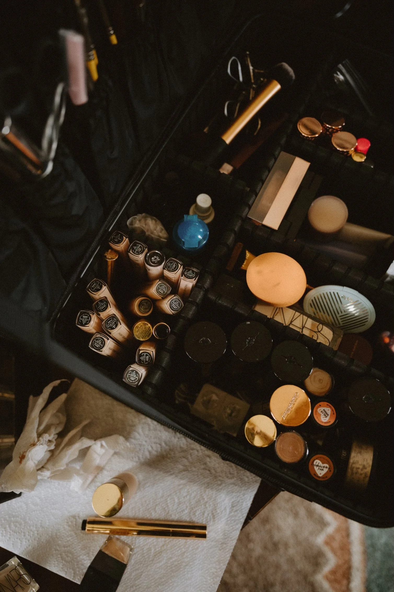 Beauty makeup products and tools organized on a black case, including lipsticks, powders, brushes, and a gold-colored mascara, with some items on a white textured cloth surface.