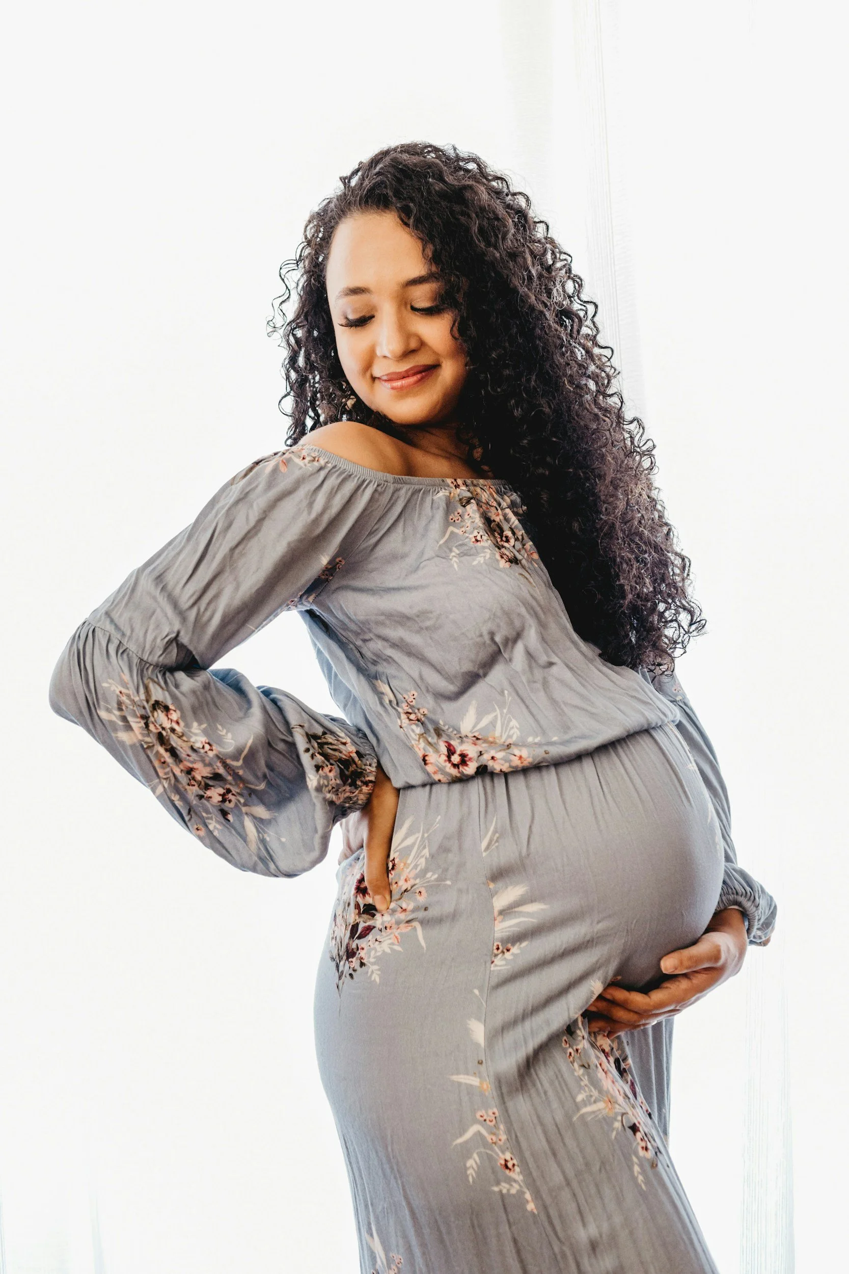 Pregnant woman with curly hair wearing a grey floral dress, holding her belly with one hand and her shoulder with the other, smiling with her eyes closed.
