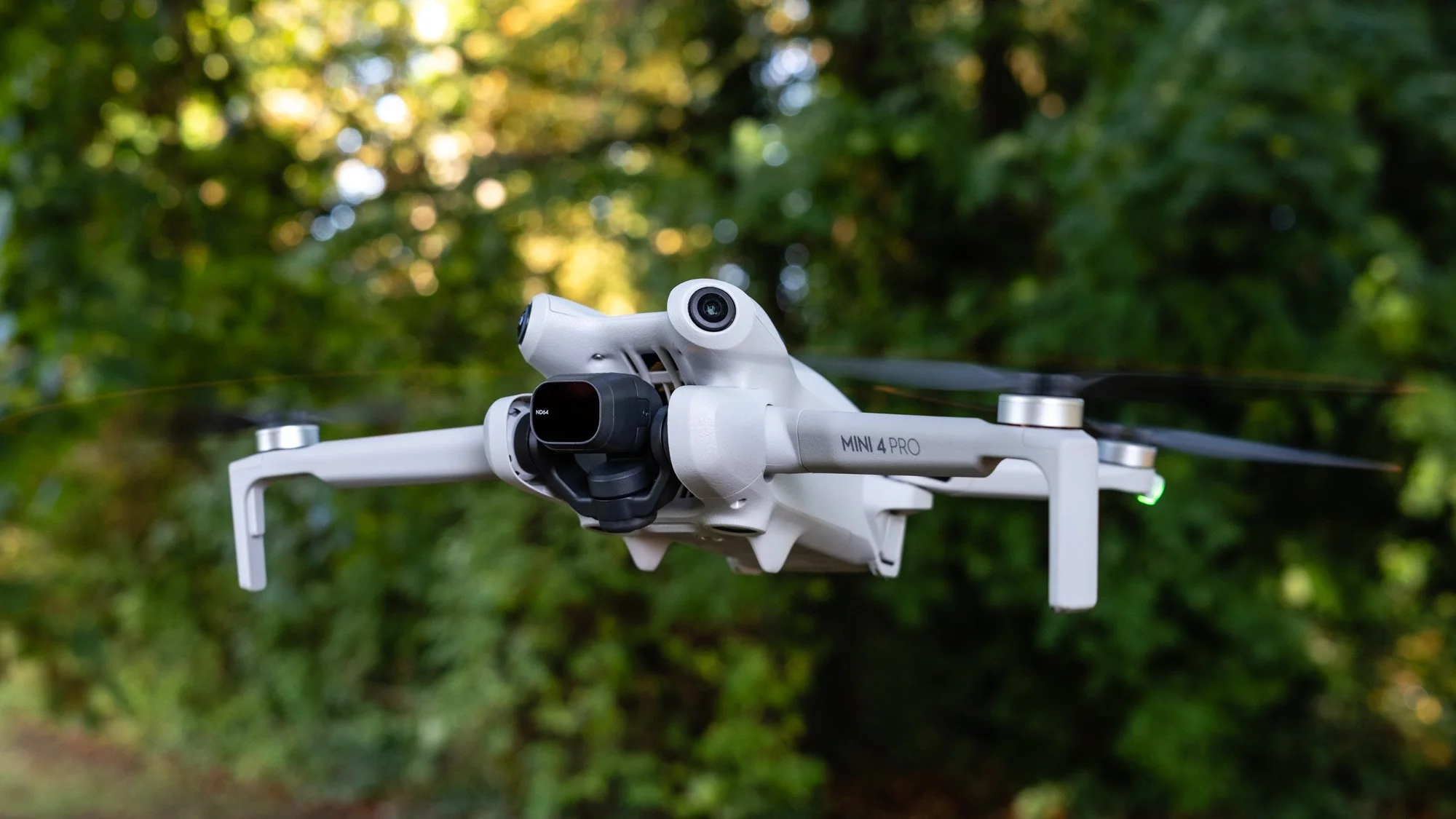 A DJI Mini 4 Pro drone in flight outdoors with a blurred green wooded background.