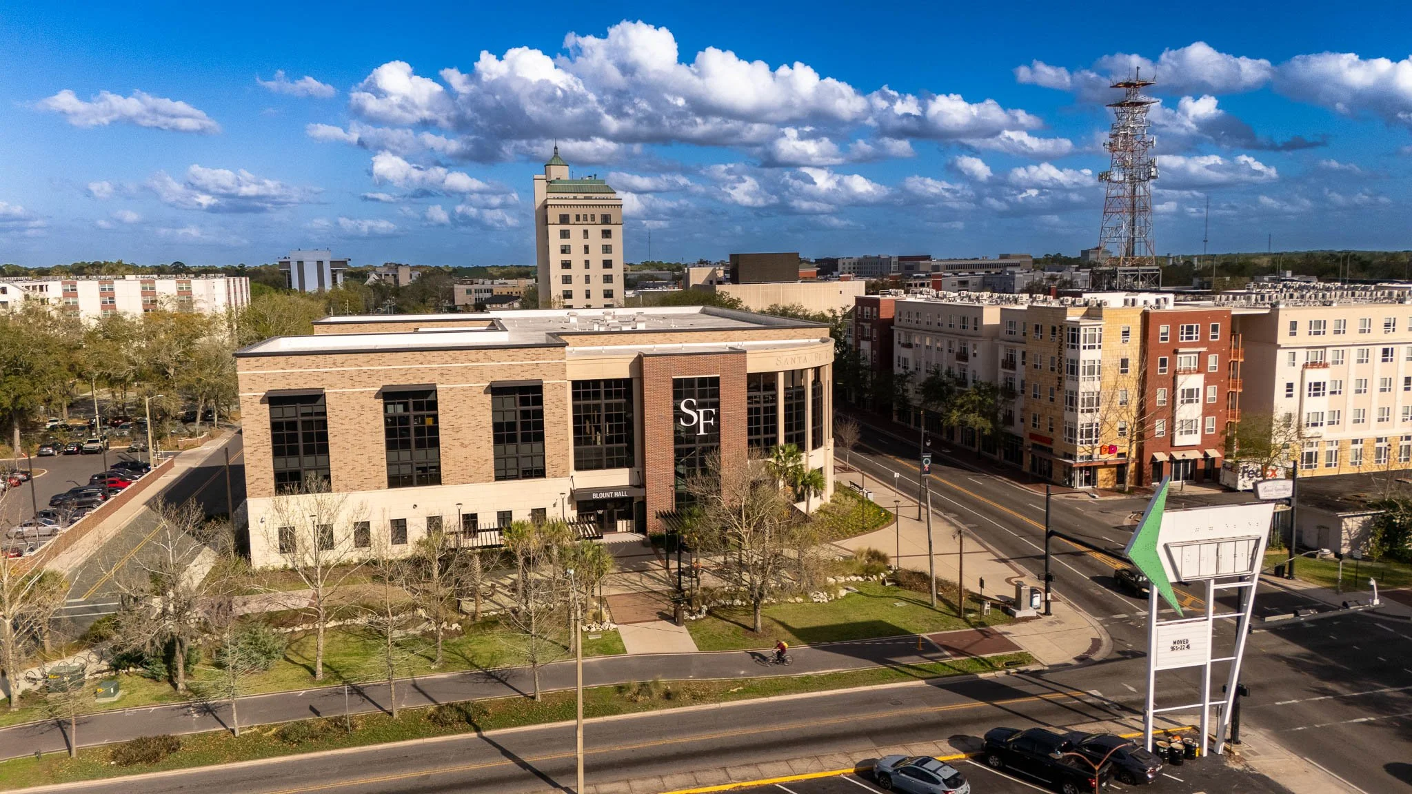 An aerial view of Santa Fe College's Charles L. Blount Center, surrounded by trees, streets, and parked cars, with a communication tower and other buildings in the background, under a partly cloudy sky.