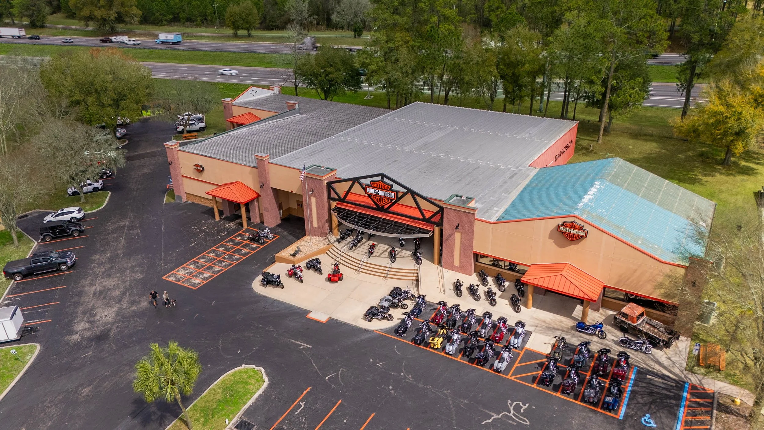 Aerial view of a Harley-Davidson motorcycle dealership with motorcycles parked in front and a large logo sign above the entrance. The building has a beige exterior with red accents, and a parking lot with several cars and a motorcycle. Trees surround the area, and there is a highway in the background.