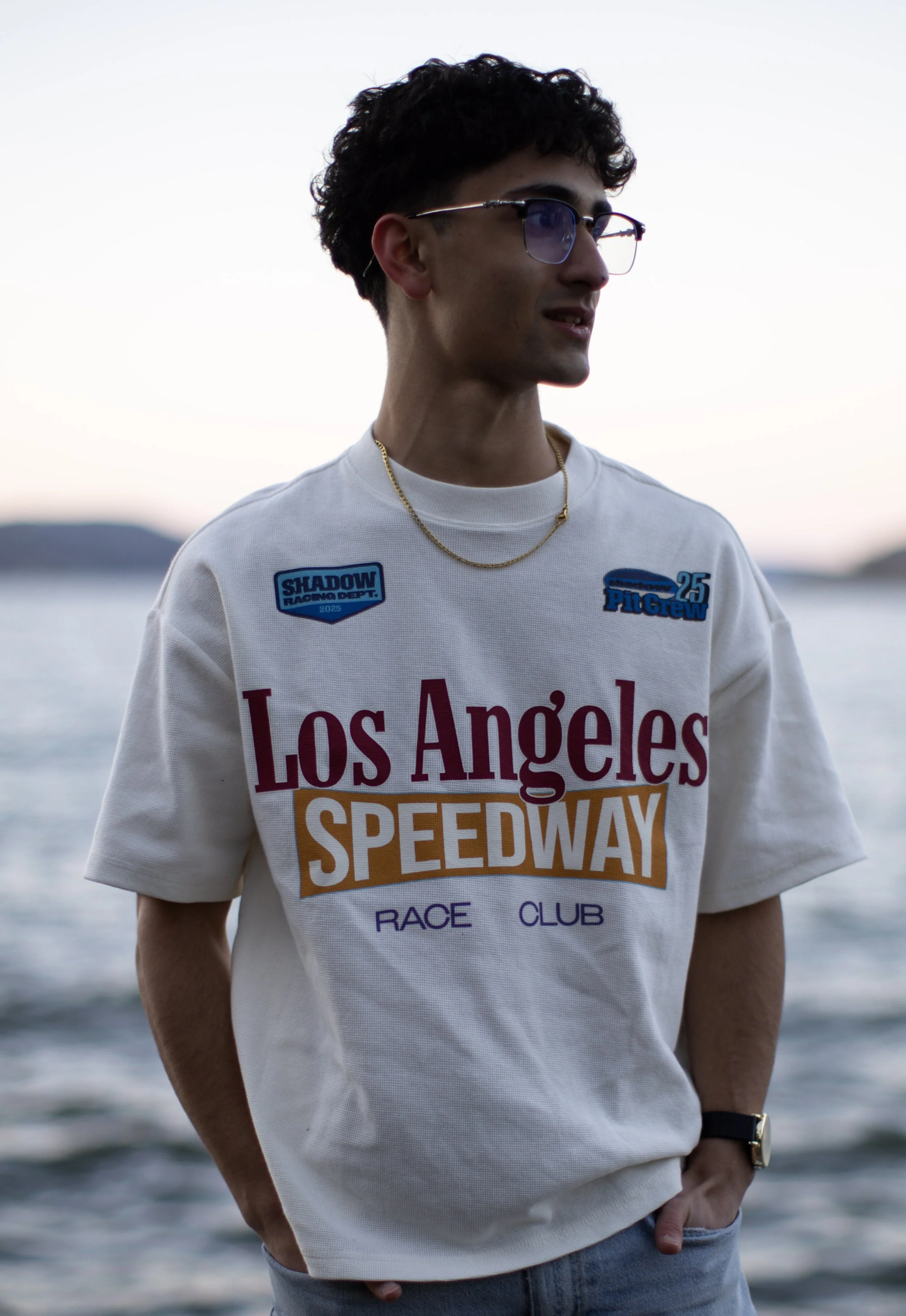 Young man wearing glasses, a white Los Angeles Speedway T-shirt, jeans, a gold chain, and a black watch standing near a body of water at sunset.