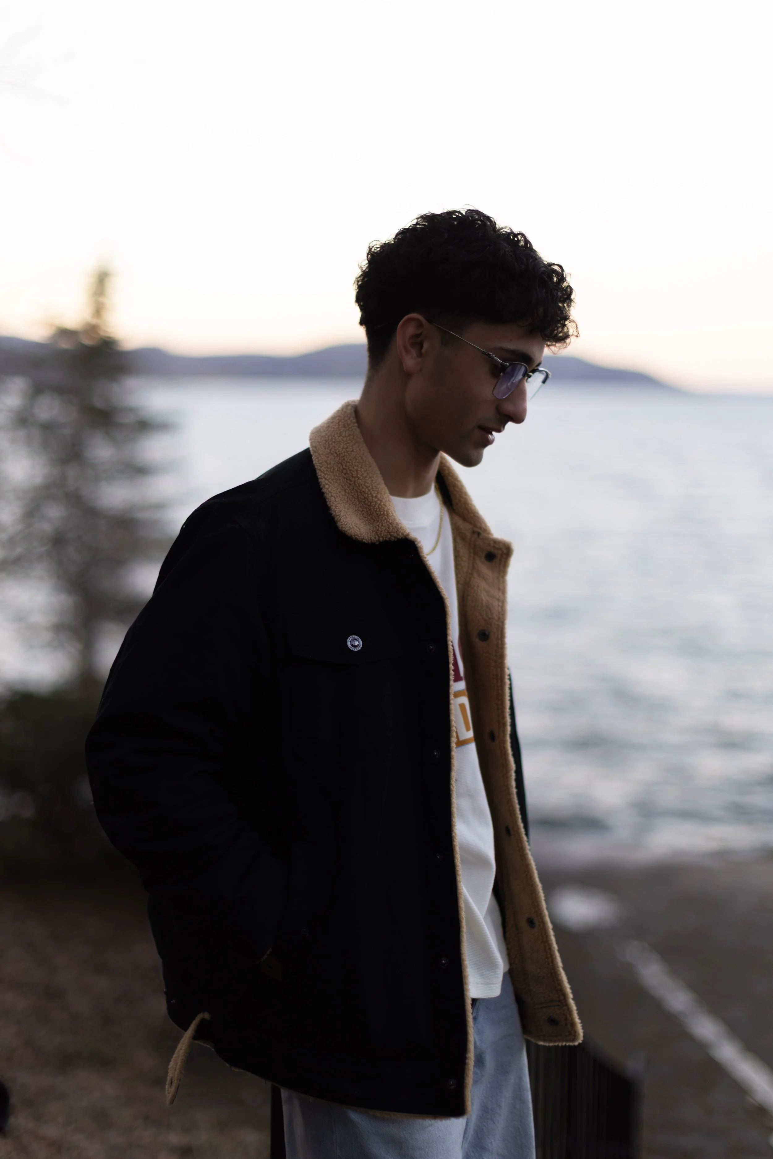 A young man with curly dark hair and sunglasses standing outdoors near a body of water during sunset, wearing a black jacket with a beige fleece lining and light-colored pants.