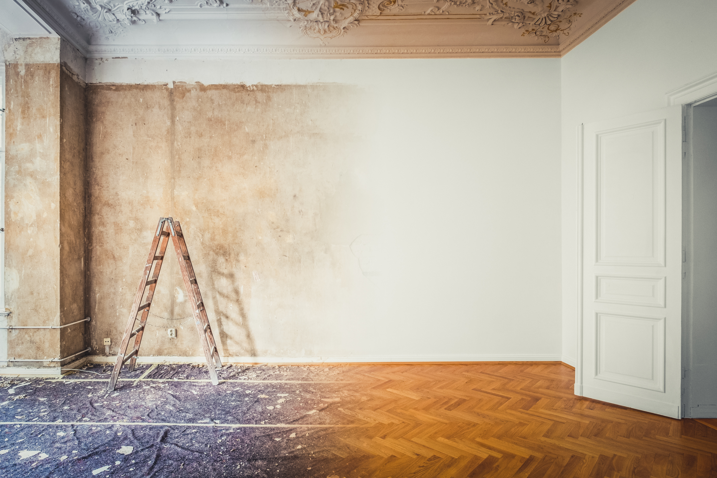 Room under renovation with a ladder, showing a partially stripped wall and a wooden floor.