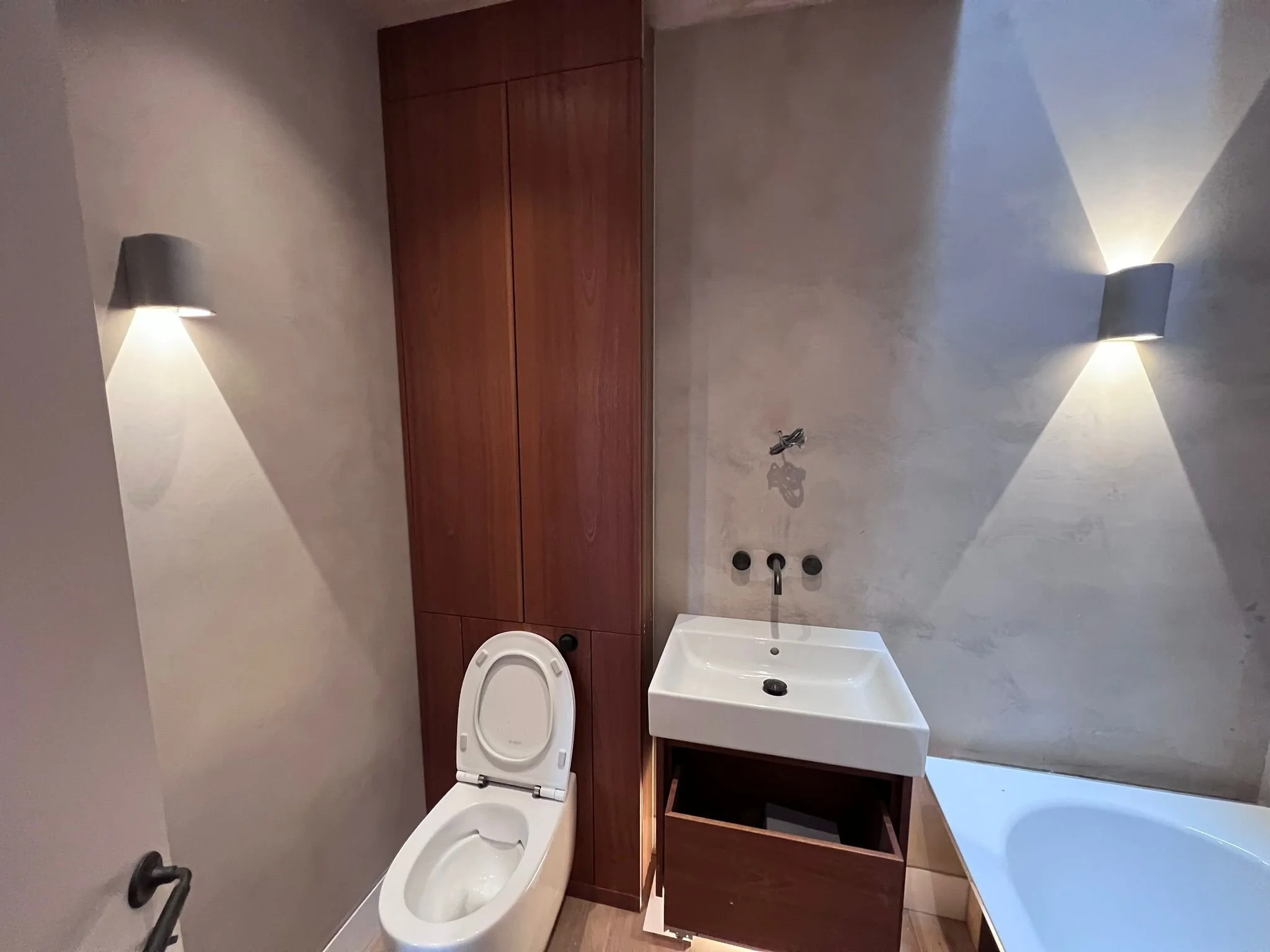 Modern bathroom with wooden cabinets, a white toilet, a white sink with a dark wooden cabinet below, and a bathtub. Wall-mounted lights illuminate the space.
