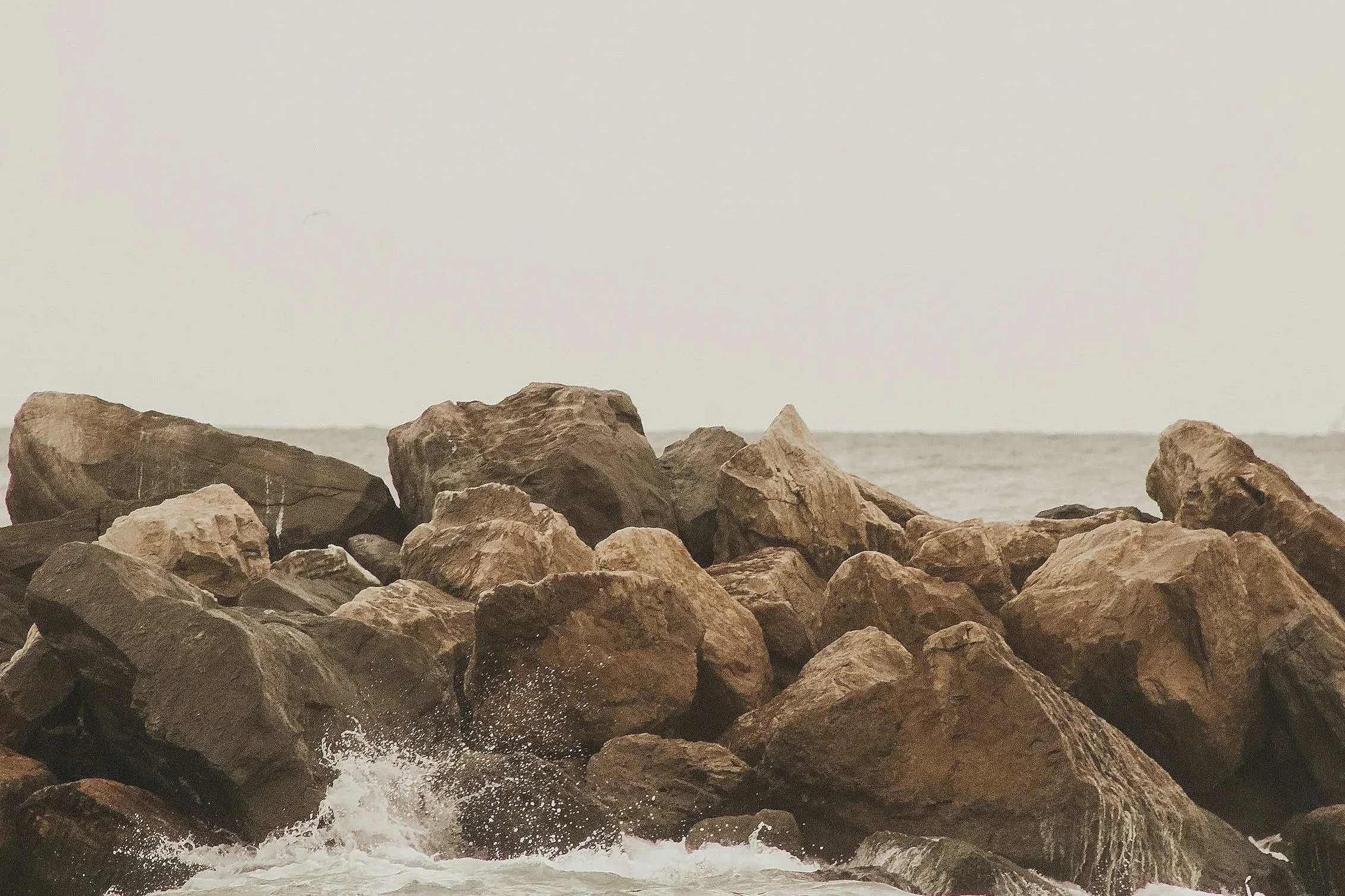 Rocks on a shoreline with waves splashing at the base, overcast sky in the background. This image demonstrates the solid ground faith gives as you battle intrusive thoughts, OCD, anxiety.