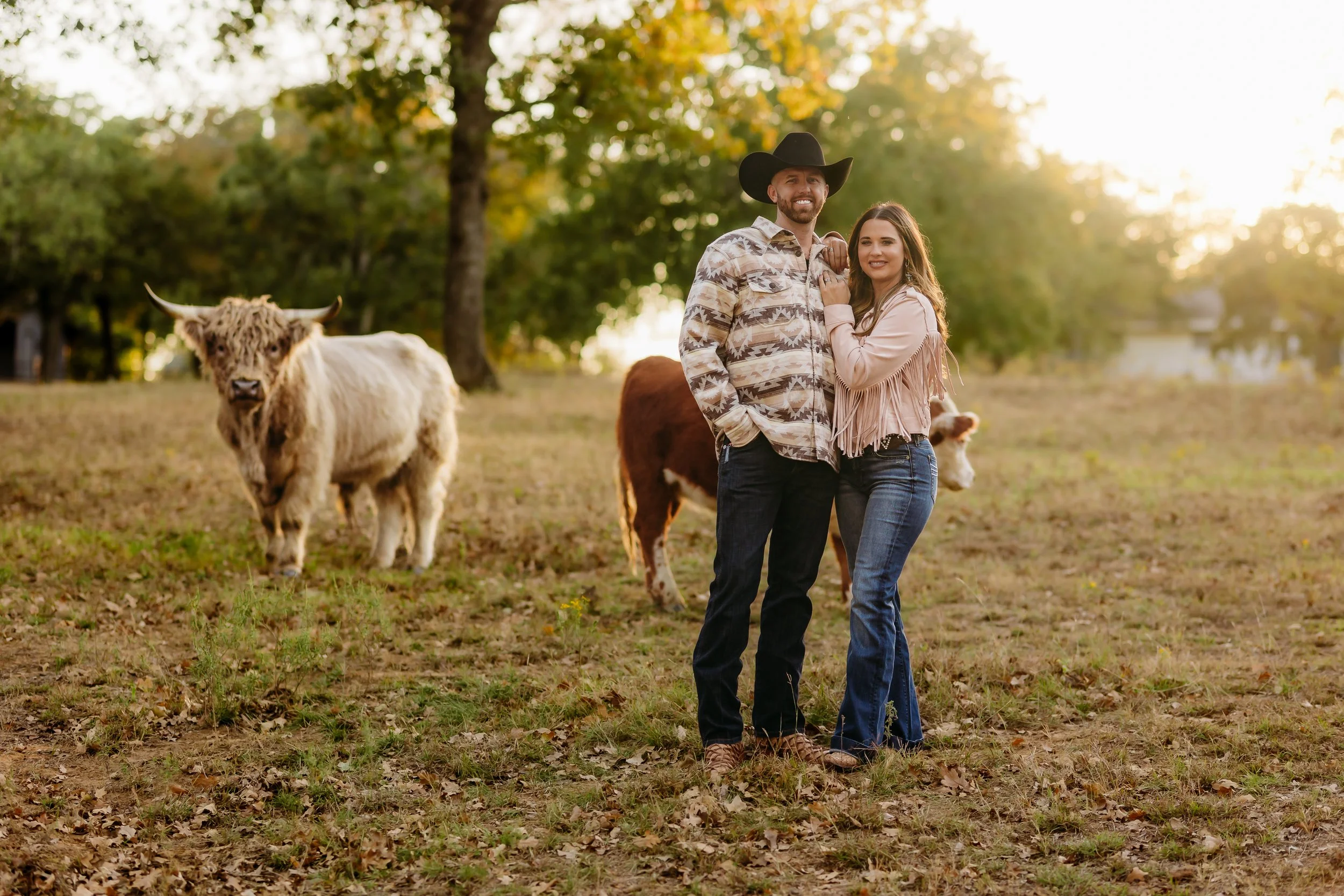 A couple standing together in a field with cows, trees, and the sun setting in the background.