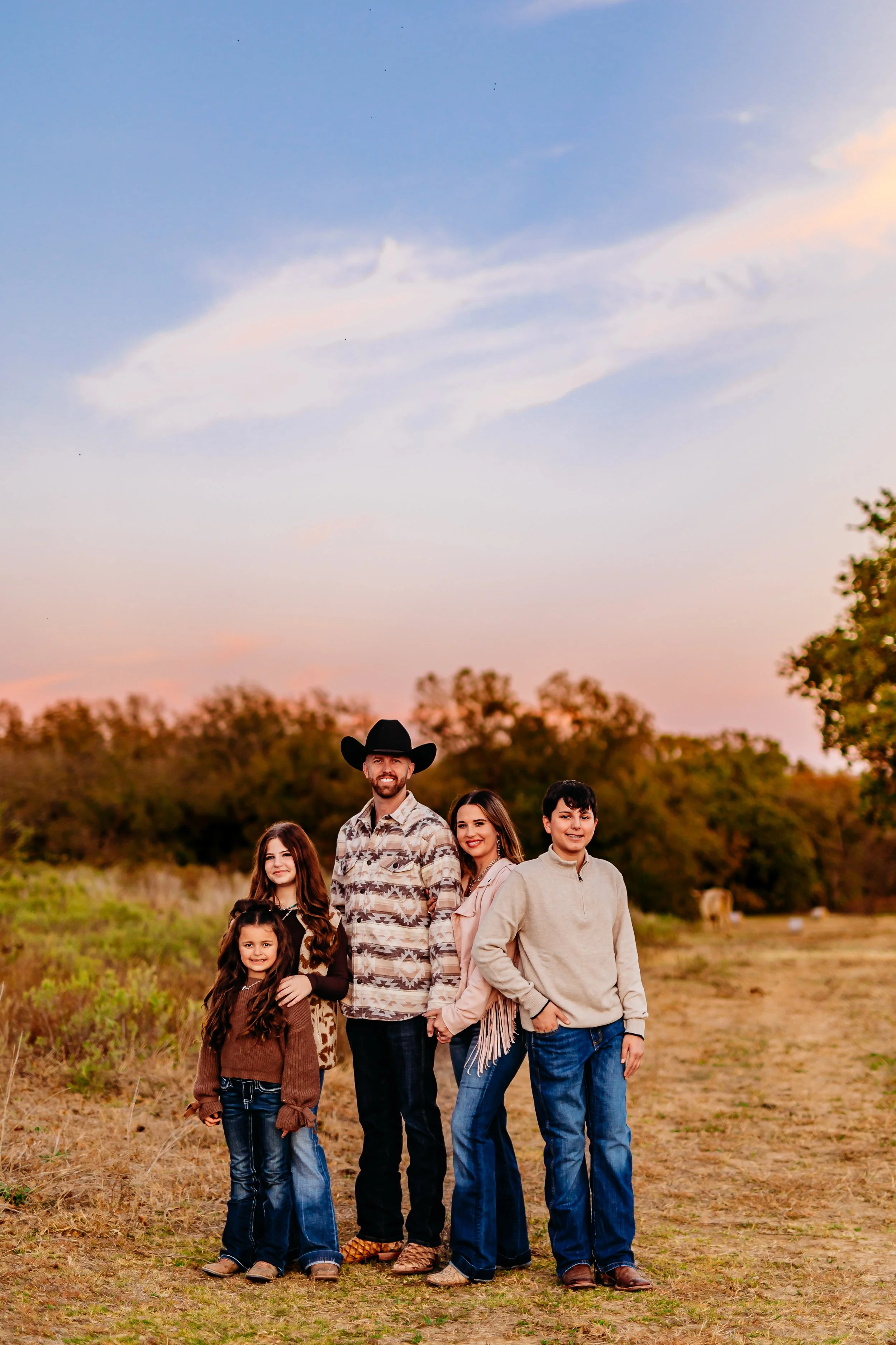 A family of five standing outdoors in a field during sunset, with trees in the background.