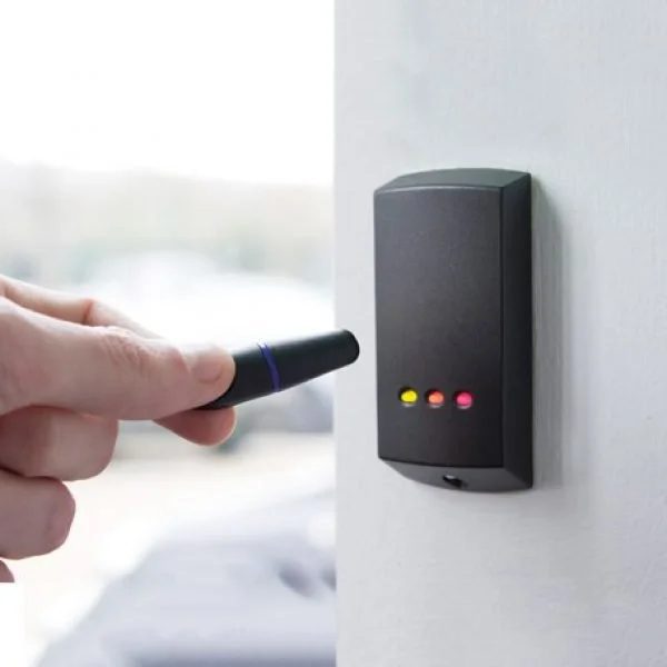 Person using a black electronic vape or vaporizer near an electronic device on a white wall with three indicator lights (yellow, orange, pink) shown.