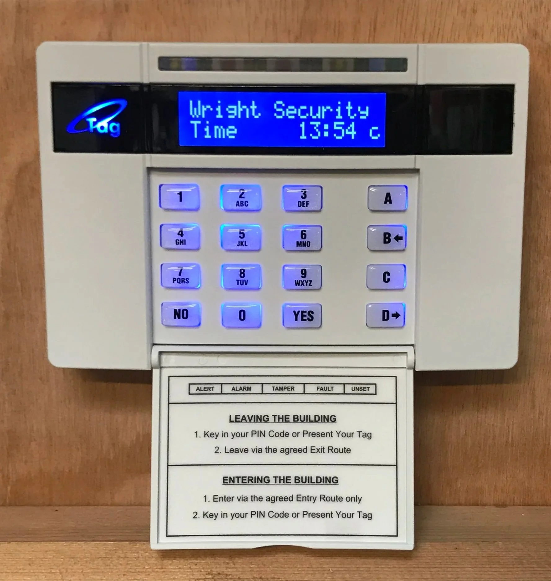 An electronic security keypad with backlit buttons, a blue screen displaying 'Wright Security' and the time 13:54, mounted on a wooden wall, with instructions for building entry and exit routes attached below.