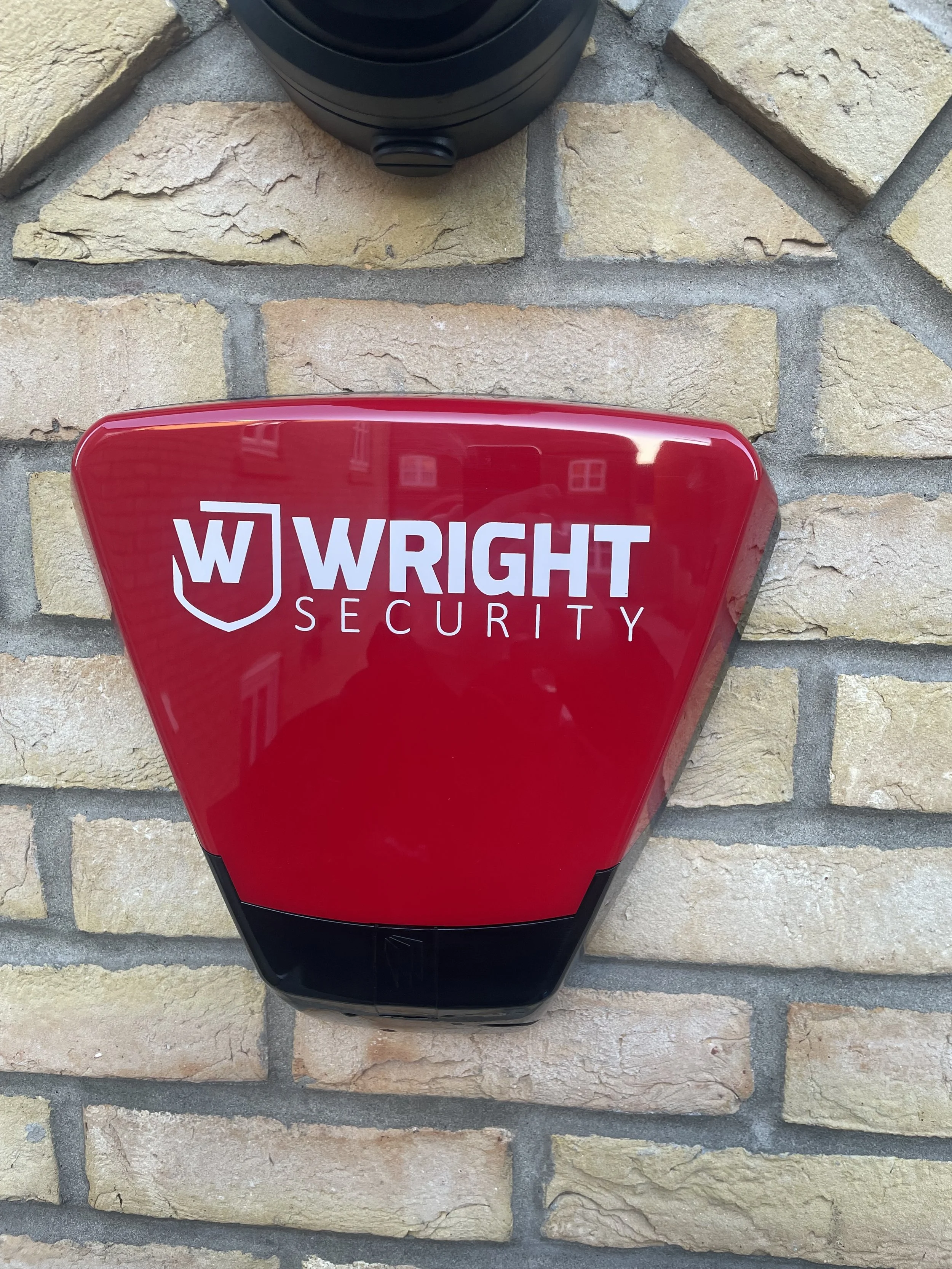 Security alarm device with red cover displaying the logo and text 'Wright Security' mounted on a brick wall.