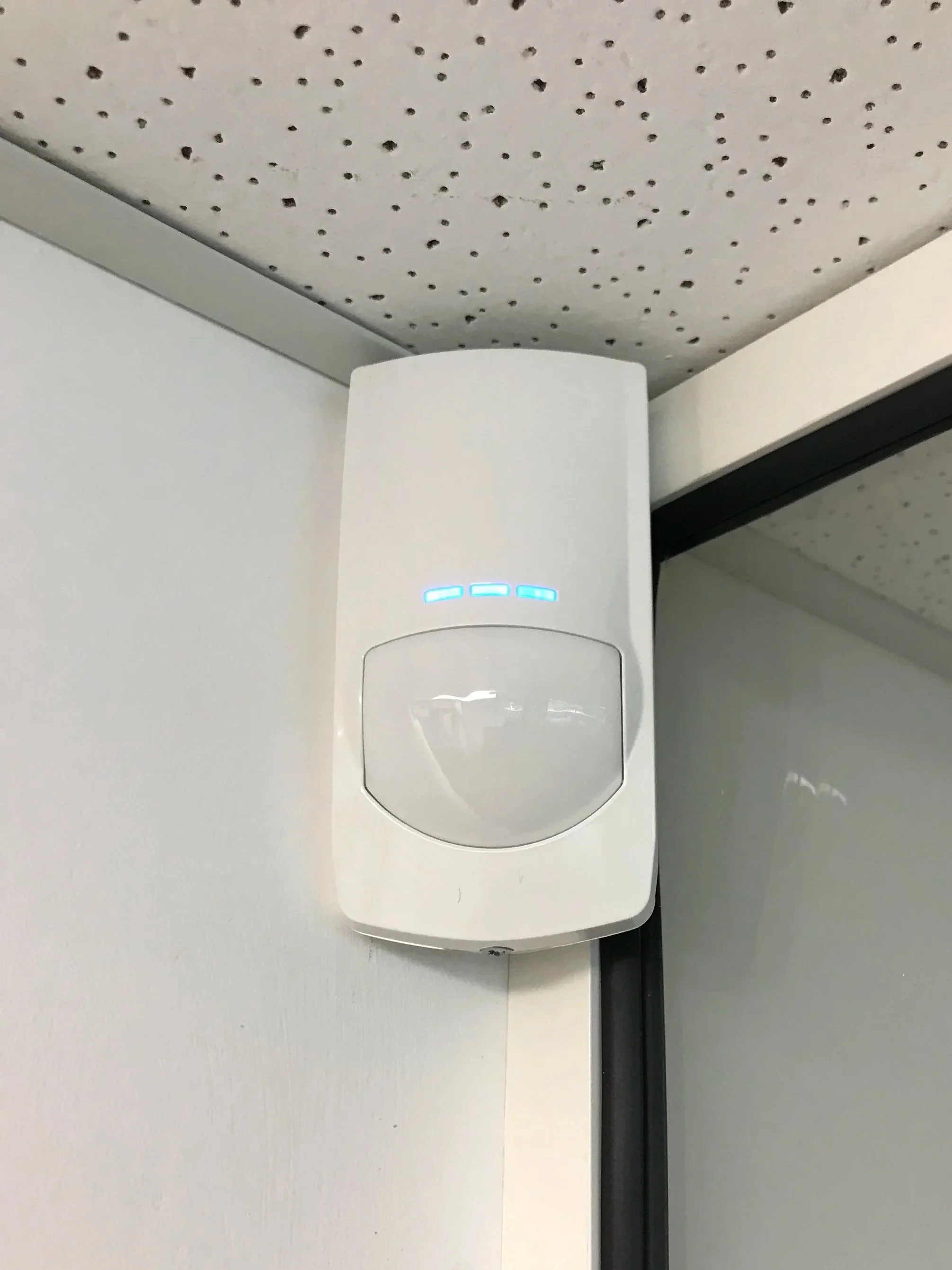 White motion sensor device with blue indicator lights mounted on a wall near the corner of a room, with a perforated ceiling above.
