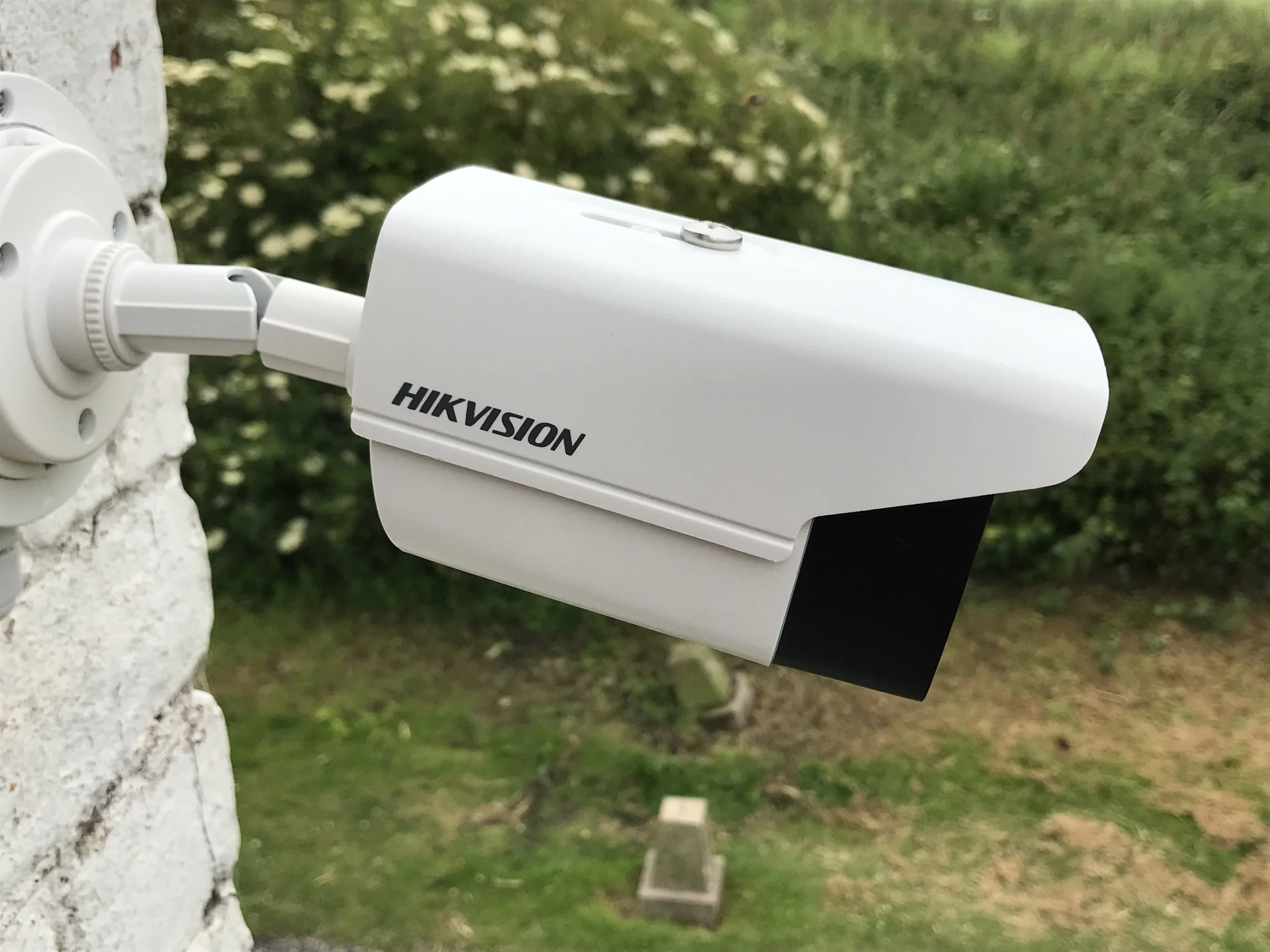 A white Hikvision security camera mounted outdoors on a white brick wall, with a background of green trees and grass.