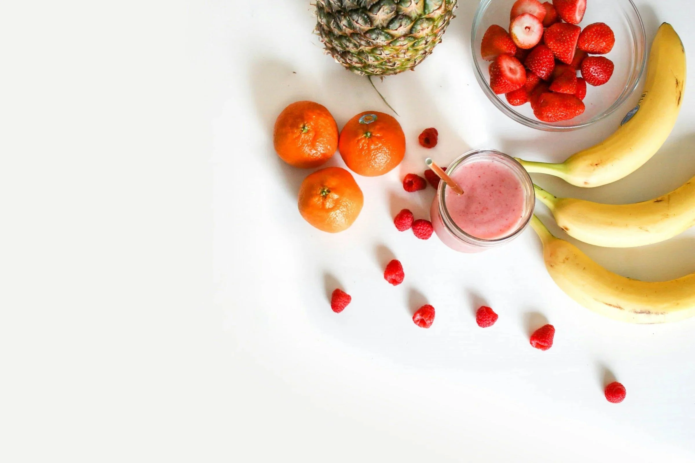 Fresh pineapple, strawberries in a glass bowl, bananas, oranges, raspberries, and a pink smoothie in a jar on a white surface.