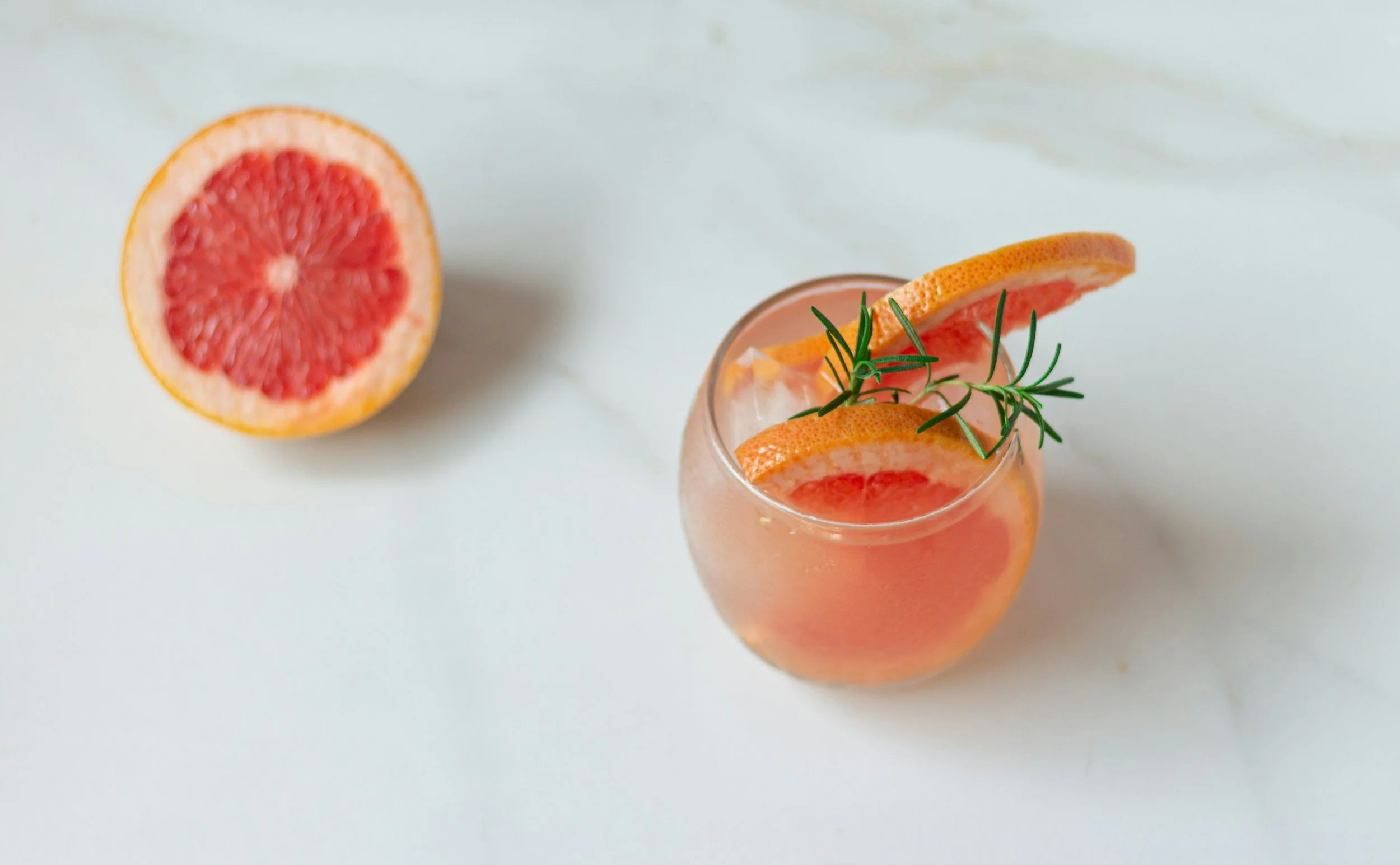 A glass of pink grapefruit cocktail garnished with a grapefruit slice and a sprig of rosemary, with half a grapefruit on a white surface in the background.