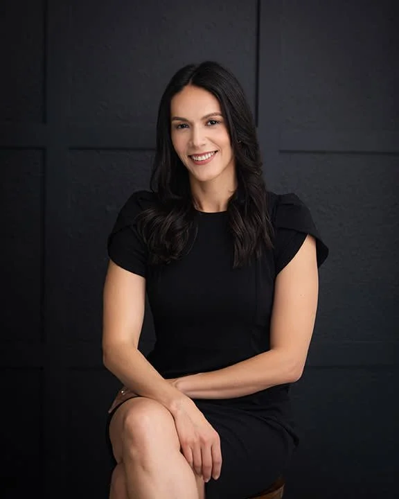 A woman with long dark hair wearing a black dress, sitting and smiling against a black textured background.