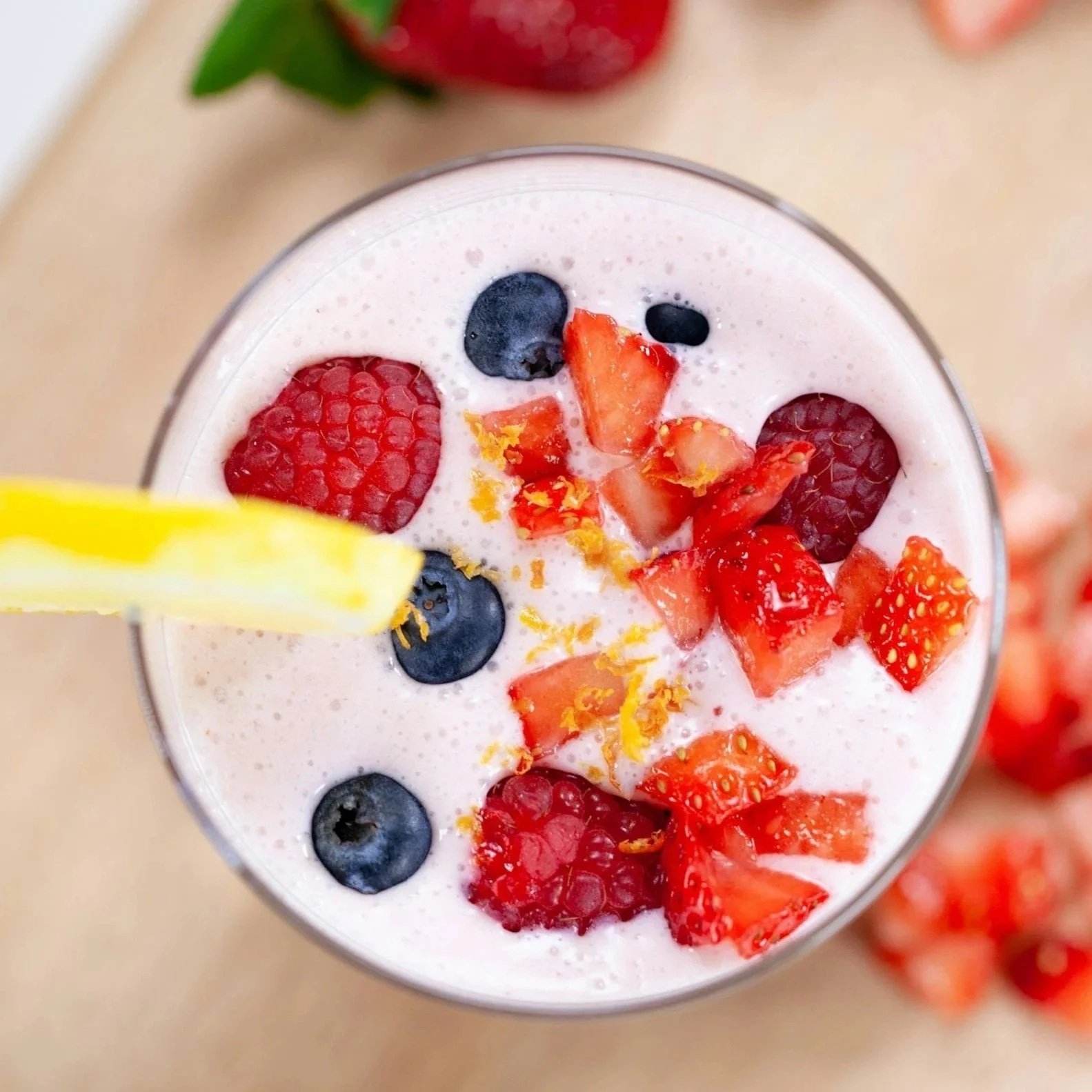 Close-up of a glass filled with a pink smoothie topped with fresh strawberries, blueberries, and orange zest, with a yellow straw on the side.