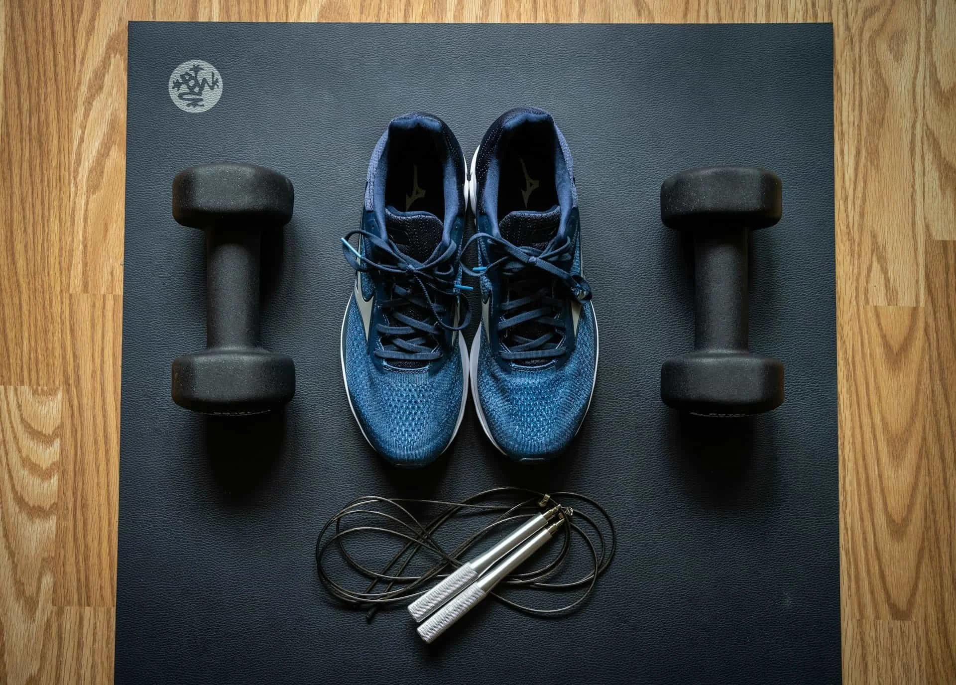 A black yoga mat with a pair of blue athletic shoes, two black dumbbells, and a black jump rope on it, on a wooden floor.