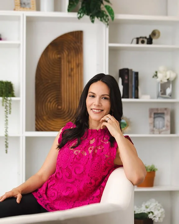 A woman with long dark hair wearing a bright pink top sitting on a white chair in a room with white bookshelves and decorative items.